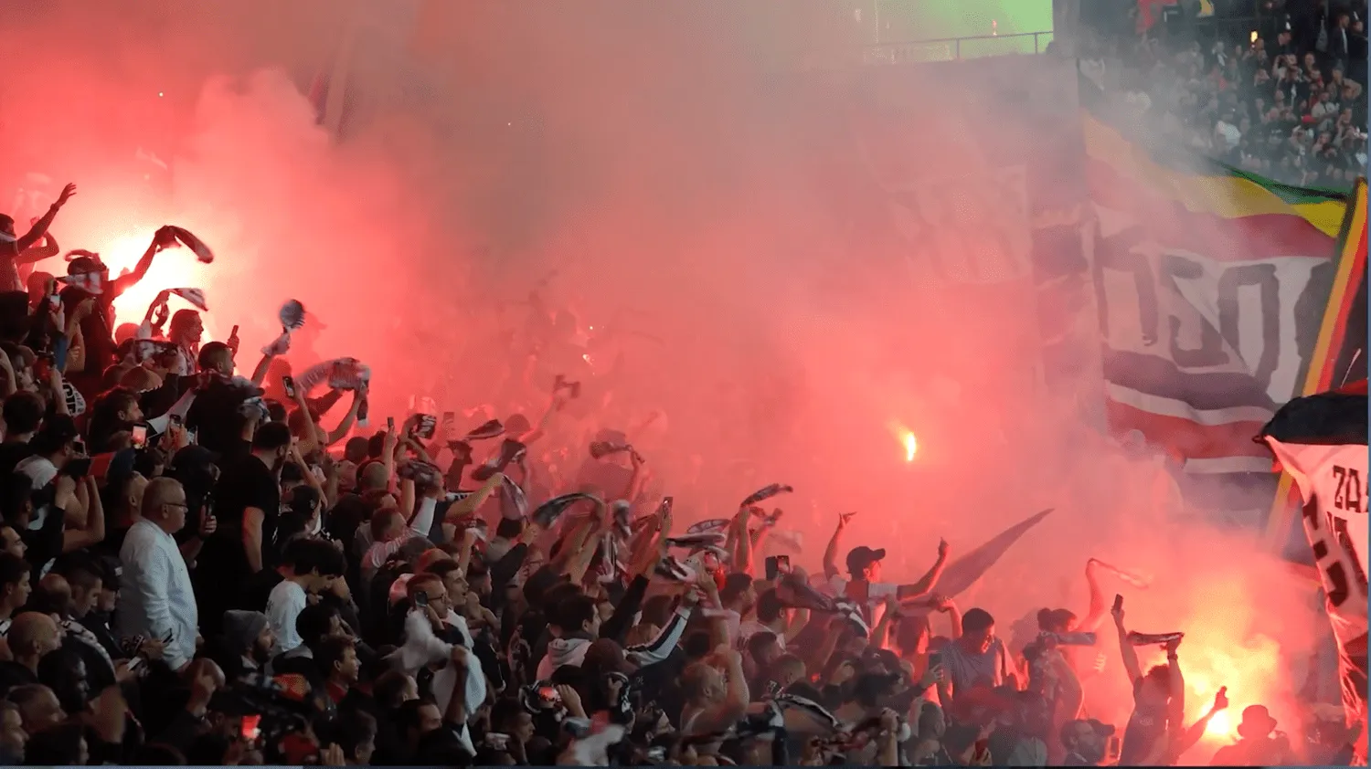 Football supporters fill a stadium with red flares, flags and celebration.